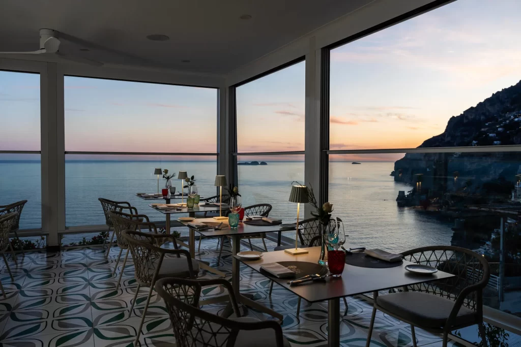Vista panoramica di un elegante ristorante con ampie vetrate sul mare al tramonto.