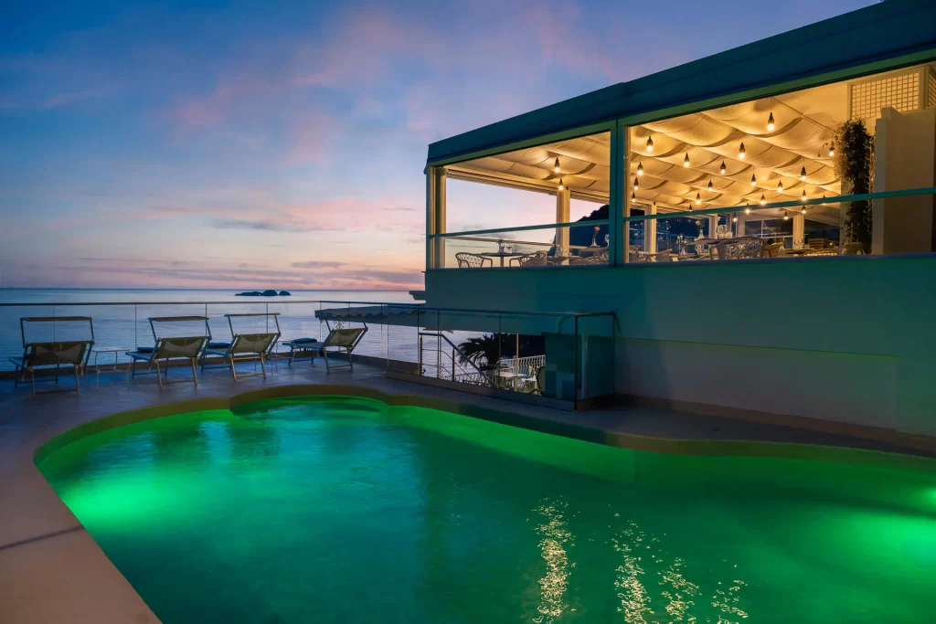 Piscina illuminata al tramonto con vista sul mare e terrazza arredata.