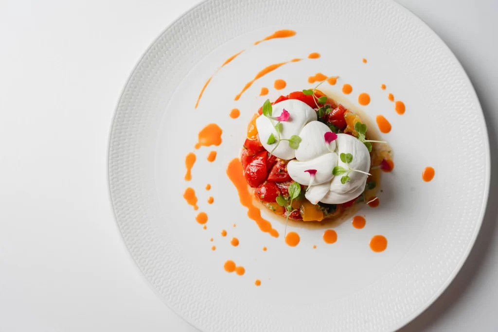 Piatto gourmet decorato con pomodori, crema e gocce di salsa su piatto bianco.