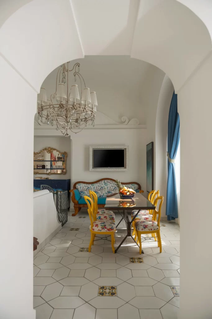Stanza luminosa con tavolo da pranzo e sedie colorate sotto un lampadario, accanto a un divano e una finestra con tende blu.