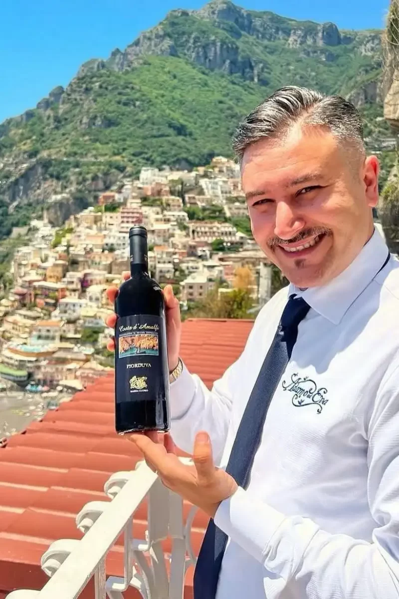 Uomo in posa con una bottiglia di vino su una terrazza con vista su una città costiera.